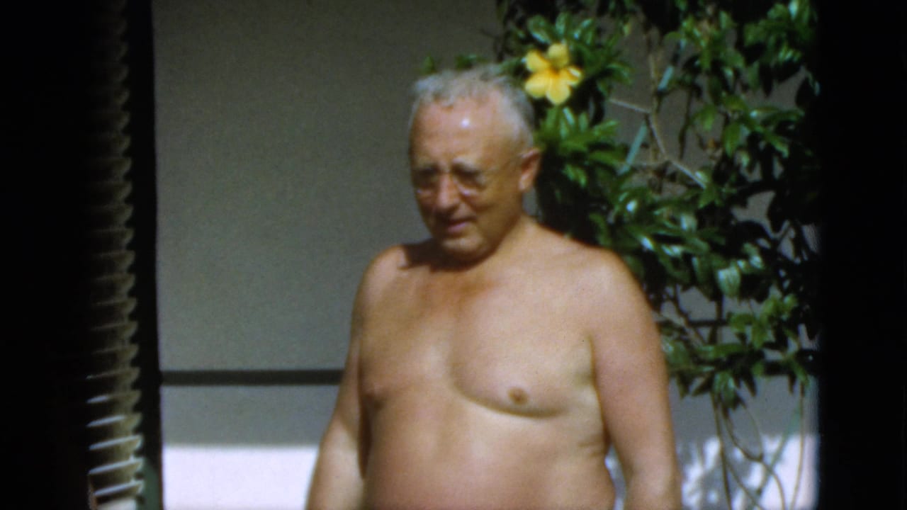 In the supplied 1952 Quebec home-movie footage, an elderly man with glasses stands shirtless in bright sun near shutters and garden foliage.