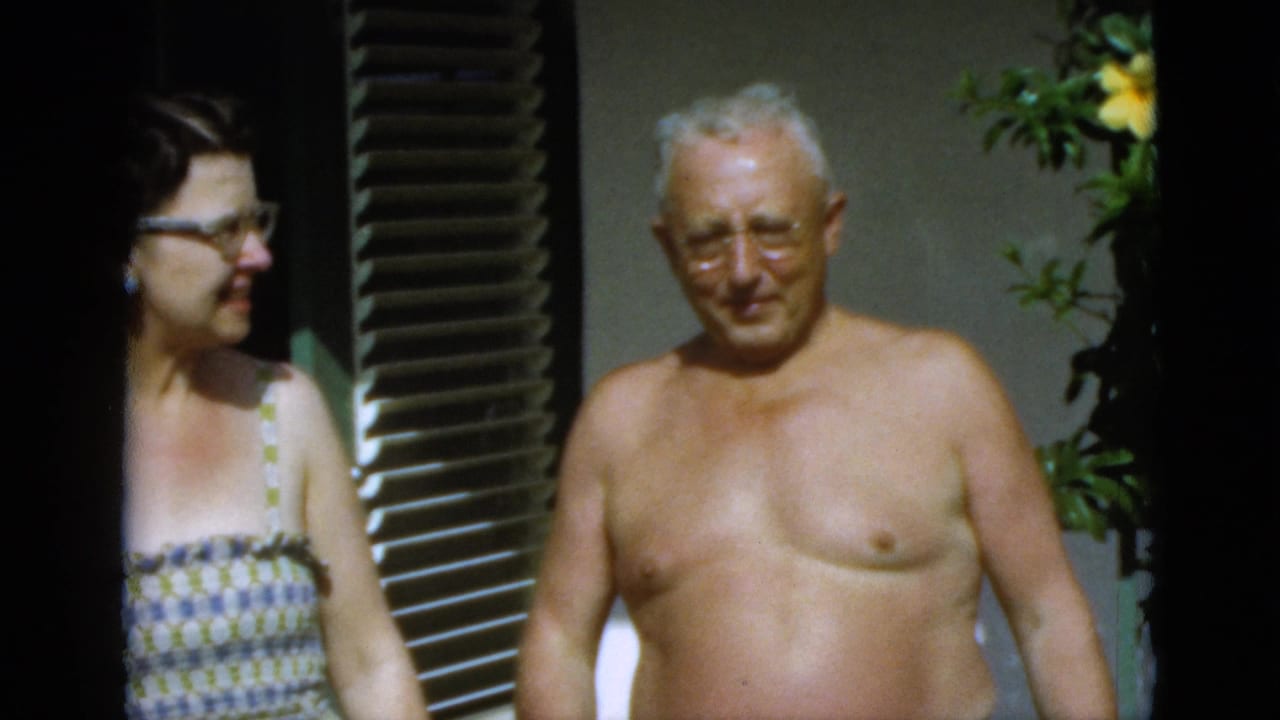 The couple appears outdoors in the supplied 1952 Quebec footage, with the woman in a patterned sundress and the man shirtless near green shutters.