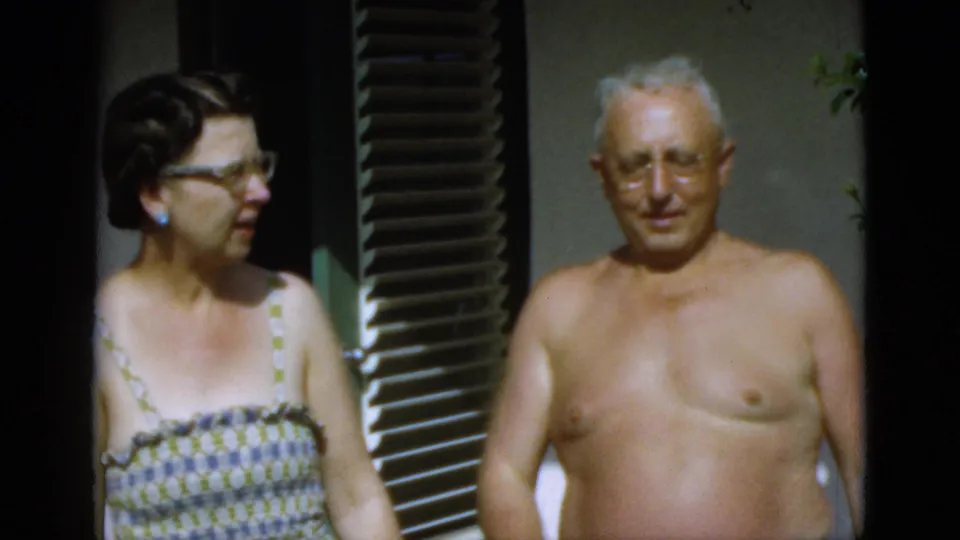 A woman in glasses and patterned sundress stands with an elderly shirtless man in bright outdoor light in the supplied 1952 Quebec footage.