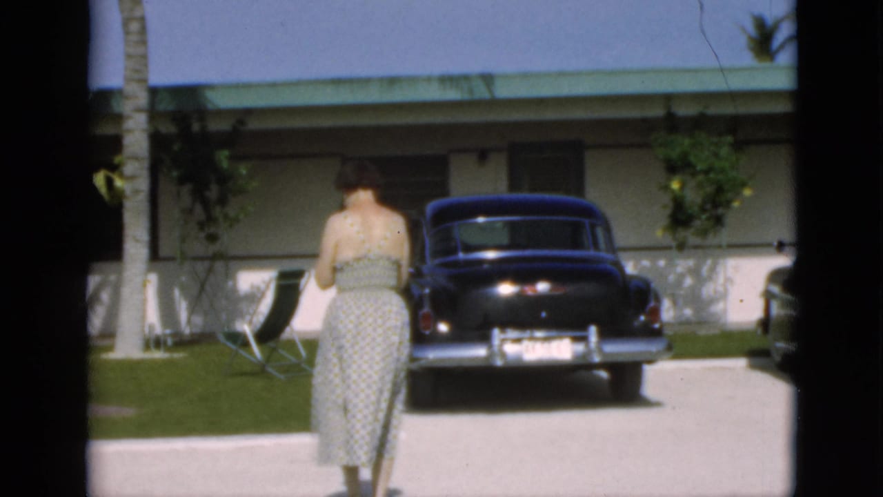 Woman in a patterned dress walks toward a dark car parked in front of a low house.