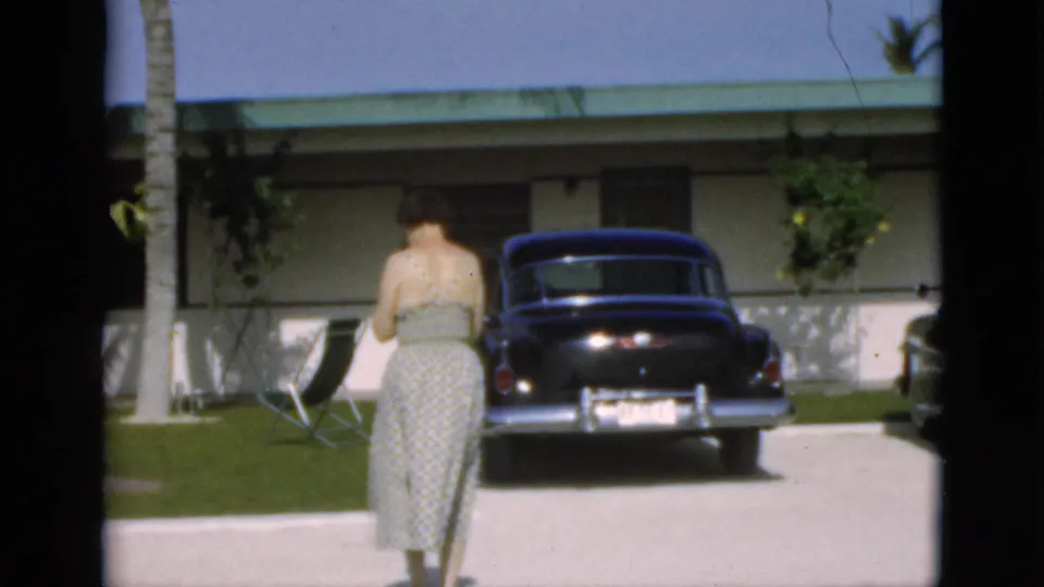 1952 8mm frame of a woman seen from behind walking across a sunlit driveway toward a dark rounded car and low house exterior.