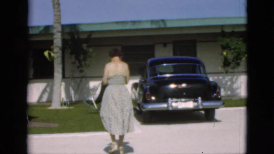 1952 8mm home-movie frame showing a woman from behind near a driveway, with a dark car, grass, palm trunk, and residential facade visible.