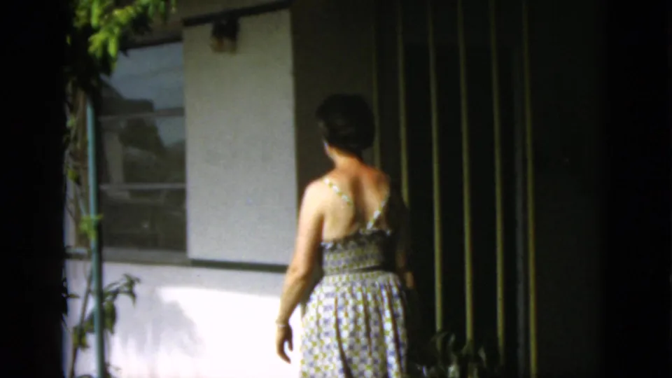 Woman seen from behind in a patterned sundress walking near a low resort-style building in metadata-identified 1952 Quebec footage.