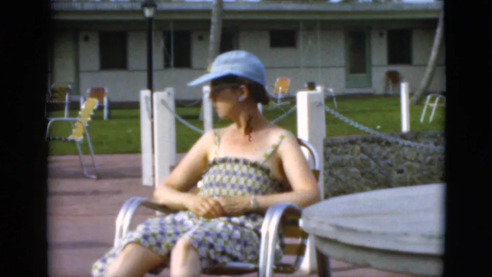 Woman in a light blue hat and patterned sundress sits in a metal patio chair at a sunny resort setting in 1952 home-movie footage.