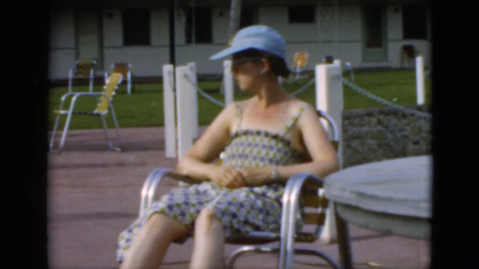 Seated woman in a patterned sundress and blue hat relaxes on a patio with lawn posts and a low building in 1952 Quebec footage.