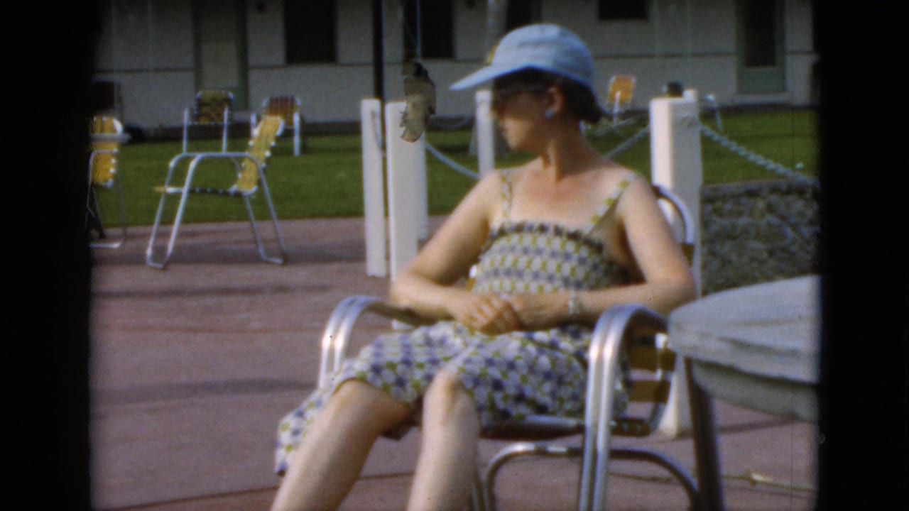In 1952 Quebec home-movie footage, a woman in a patterned sundress and light blue cap sits on a metal patio chair near yellow folding chairs.