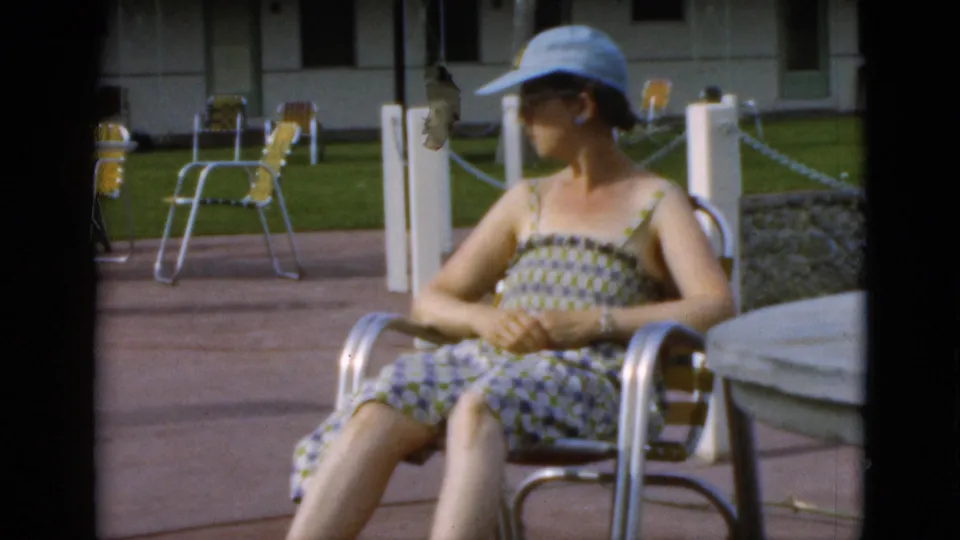 In 1952 Quebec home-movie footage, a woman in a patterned sundress and light blue cap sits on a metal patio chair near yellow folding chairs.
