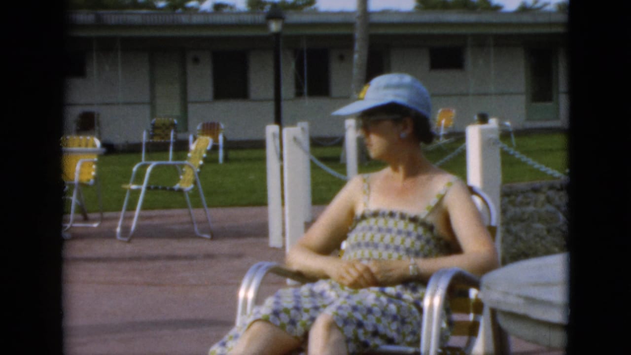 The woman remains seated on the Quebec resort patio, with a round table, white posts, lawn, and yellow folding chairs behind her.