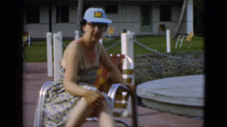 A woman in a patterned sundress and cap faces the camera from a metal chair on a paved patio at the Quebec resort setting.