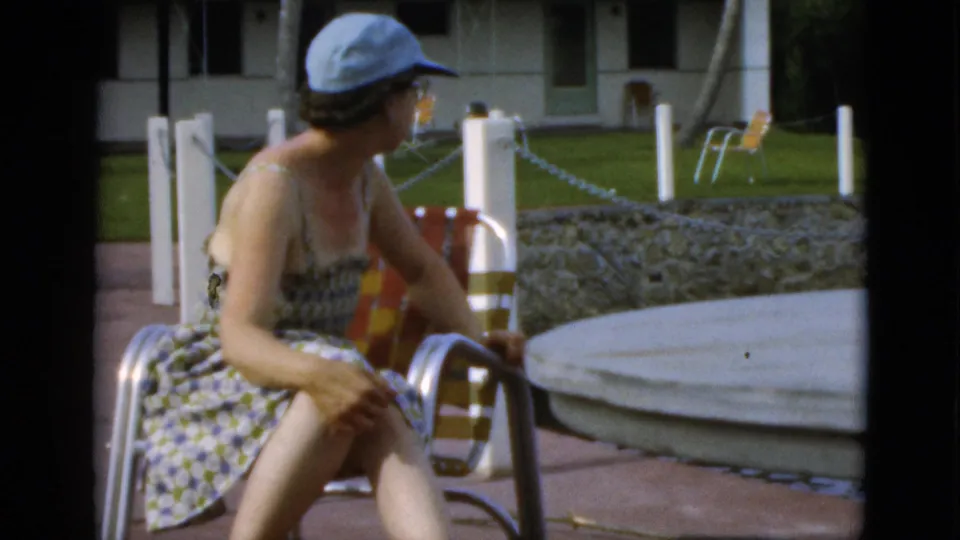 The woman turns to the side in her metal chair, with the patio table, low stone wall, white posts, lawn, and resort building visible behind her.