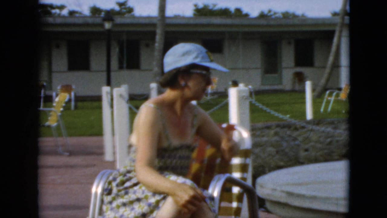 In supported 1952 Quebec footage, a woman sits outdoors in a striped lounge chair beside a patio table, lawn, posts, and low lodging rooms.