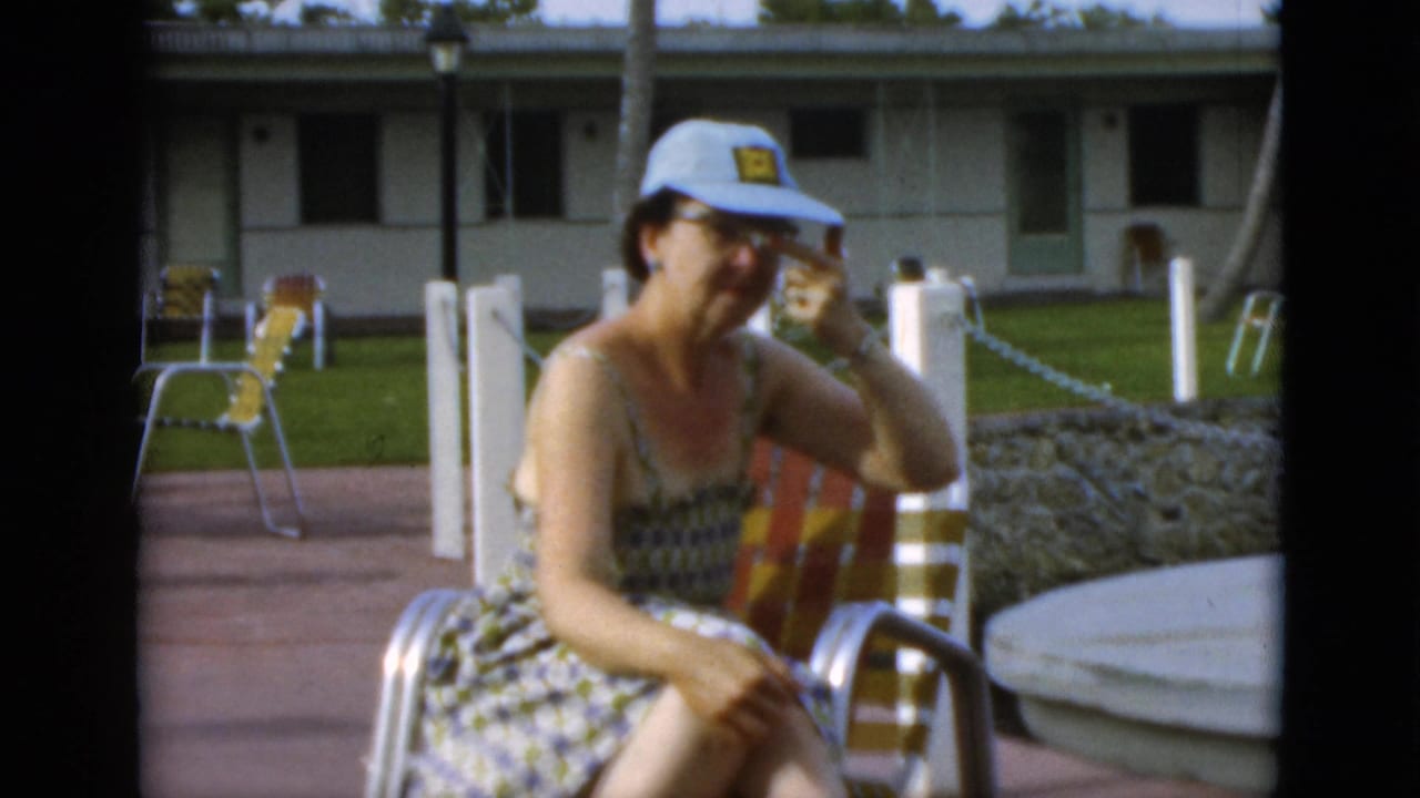 The seated woman faces the camera on a sunny patio, with a striped chair, stone planter, lawn, posts, and motel-style rooms behind her.