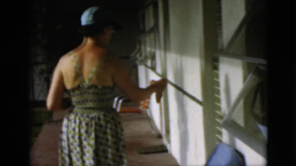 The woman walks away along a shaded porch in a patterned sundress and cap, passing white walls, open windows, and a walkway.