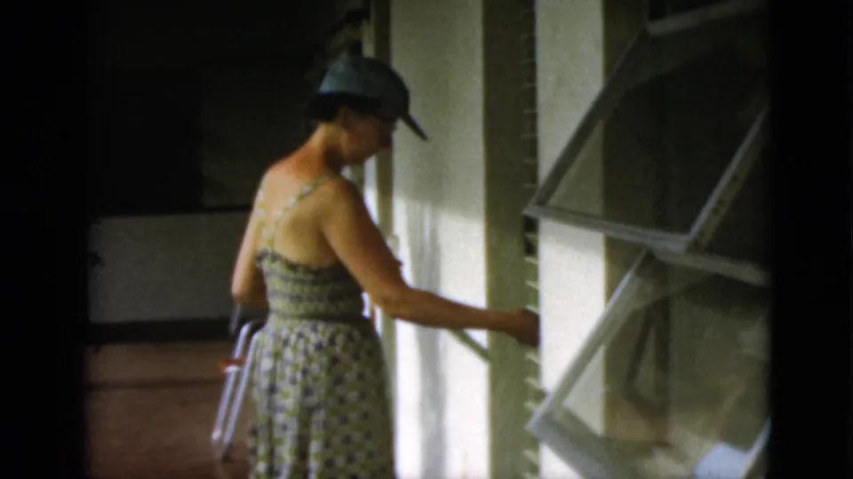 On the shaded porch, the woman reaches toward an open window shutter or panel, with white siding, porch supports, and walkway visible.
