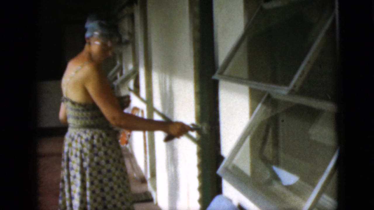 In supplied 1952 Quebec home-movie context, a woman in a patterned sundress and wide-brimmed hat opens a hinged window shutter.