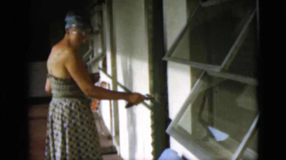 In supplied 1952 Quebec home-movie context, a woman in a patterned sundress and wide-brimmed hat opens a hinged window shutter.