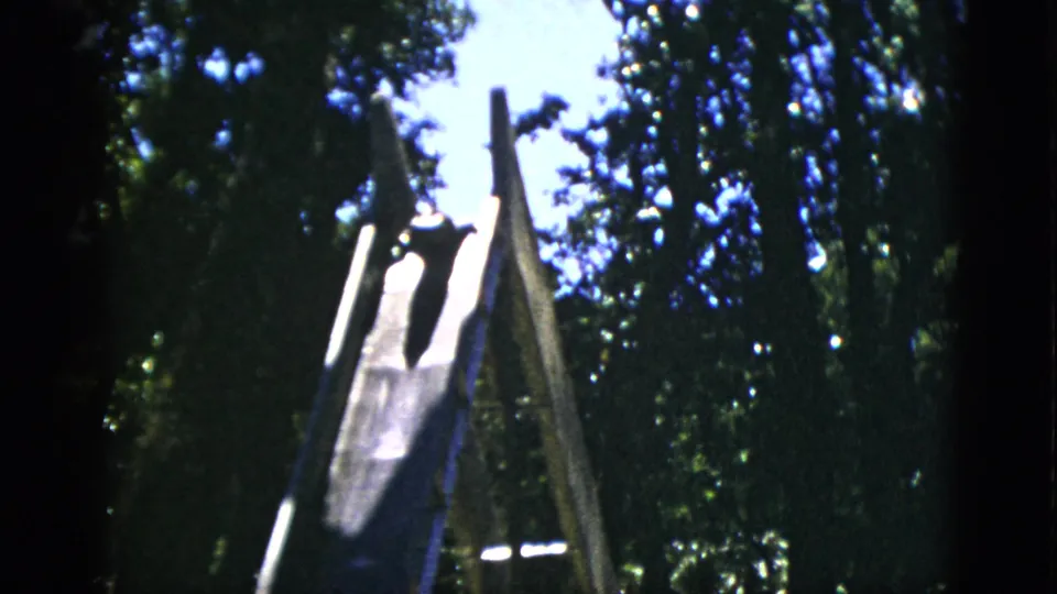 8mm home movie frame circa 1952, Quebec, Canada: wooden A-frame slide with a dark animal shape at top, framed by dense trees and bright sky.