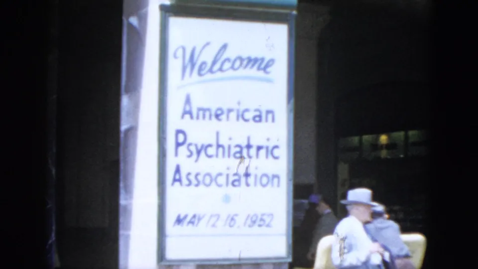 A freestanding sign reads 'Welcome — American Psychiatric Association, May 12-16, 1952'; a person in a white hat sits nearby in a building entrance area.