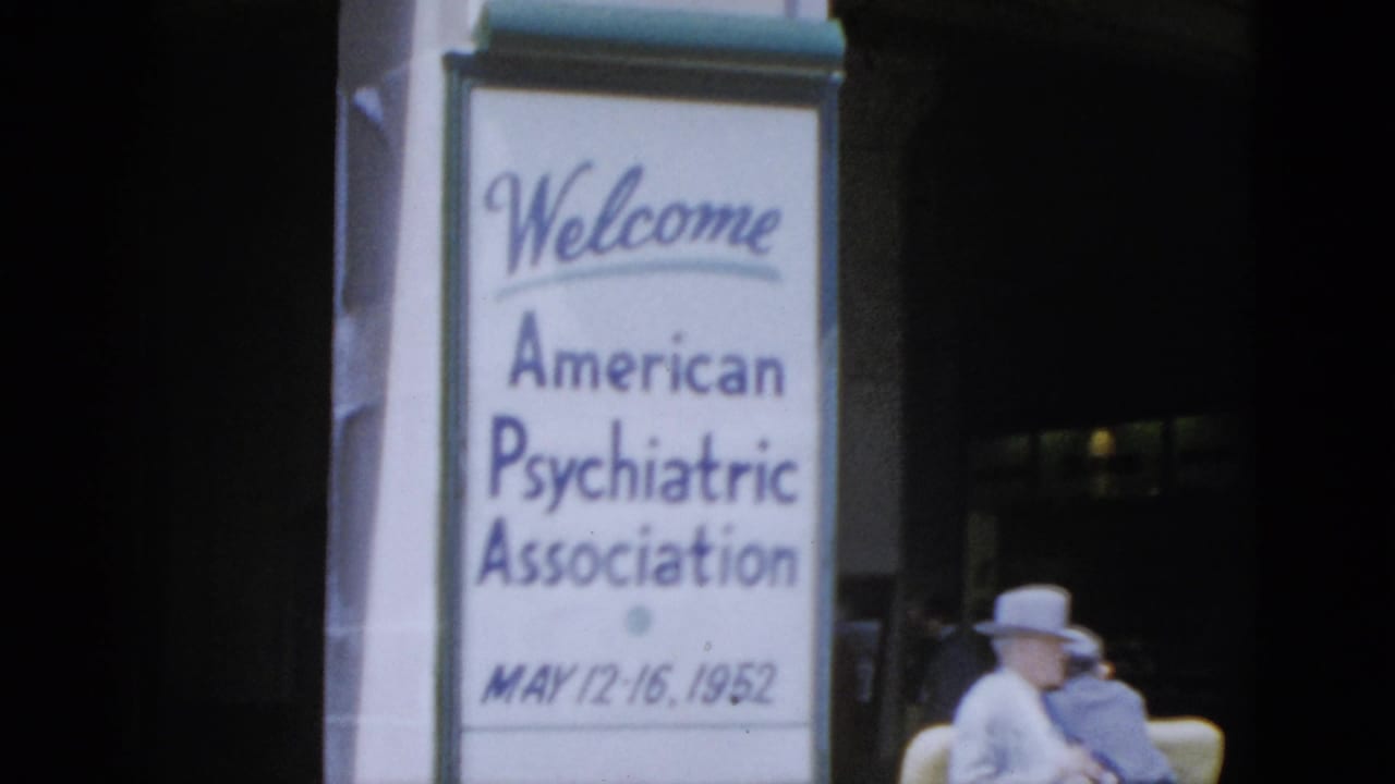Large white vertical banner reading Welcome American Psychiatric Association MAY 12-16, 1952 on a dark pillar; adult in wide-brimmed hat seated in yellow chair at right.