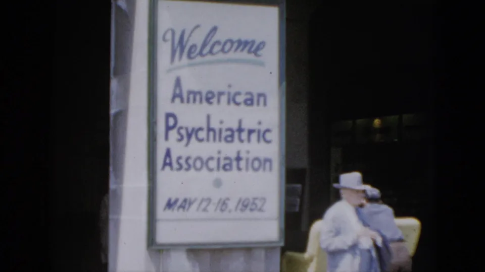 Mid-clip 8mm frame from 1952 Quebec APA conference; welcome banner legible on pillar, attendee in light suit and wide-brimmed hat seated in yellow chair to sign's right.
