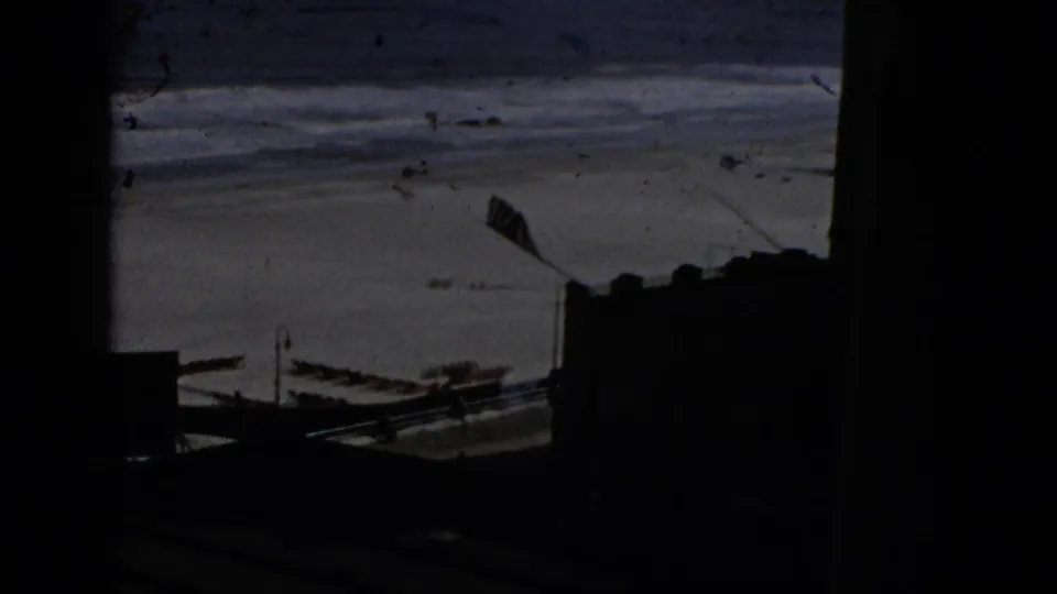 Brief 8mm exterior shot of a waterfront or harbor showing open water with waves, moored or beached small boats, and dark structural silhouettes; cooler neutral color tone.
