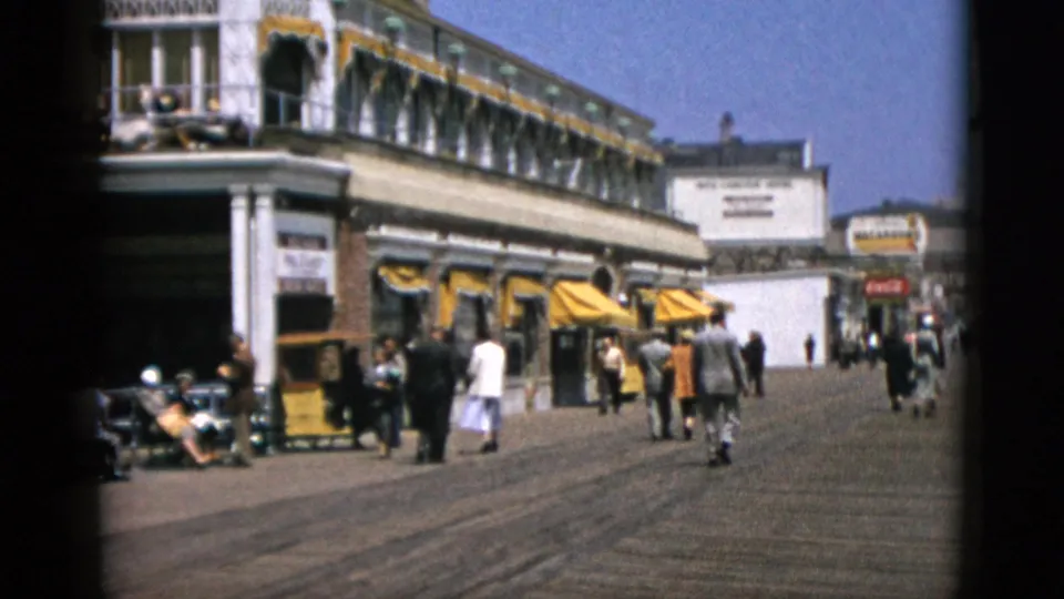 Well-lit boardwalk showing yellow-awning storefronts, period-dressed pedestrians, and a Coca-Cola sign at right, 8mm, stated Quebec, Canada, 1952.