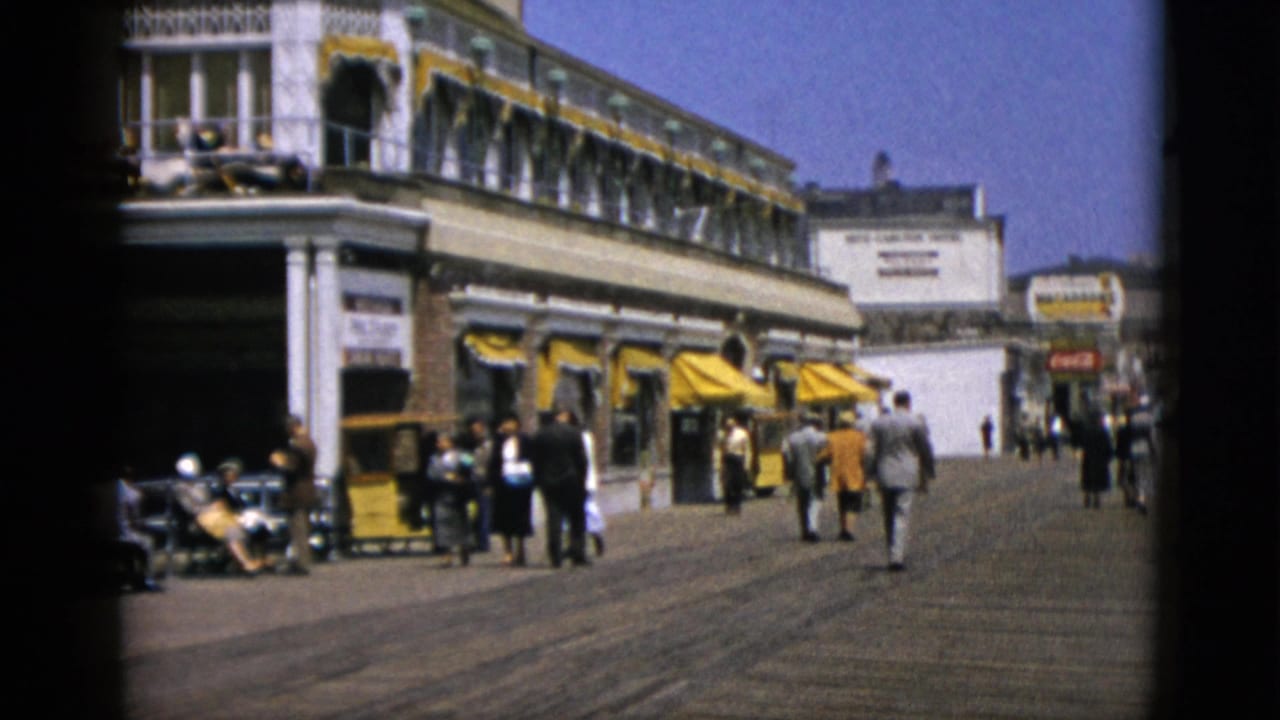 Pedestrians walking along a wide boardwalk lined with shops bearing yellow awnings, circa 1950s.