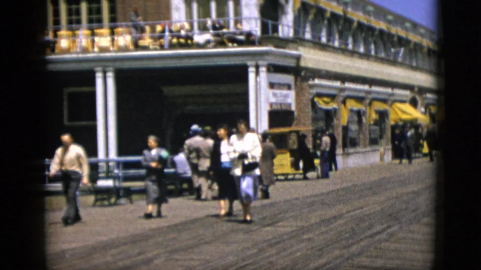 Mid-century pedestrians (women in skirts, men in trousers) on the boardwalk outside a brick building with veranda, c. 1952 Quebec, Canada.
