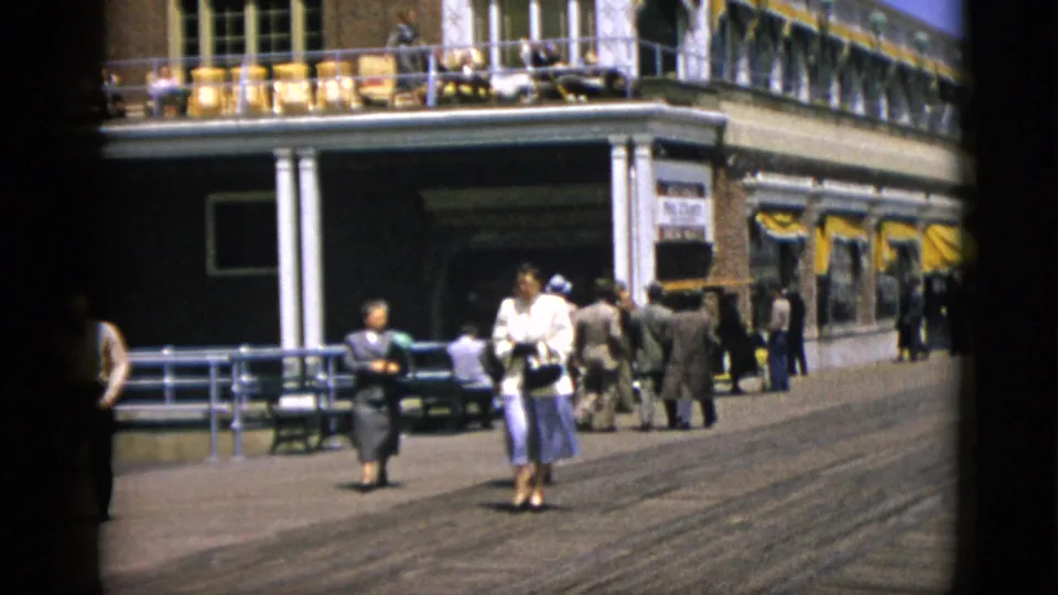 Adults in mid-century dress strolling the boardwalk, c. 1952 Quebec, Canada, with the brick commercial building and yellow awnings in the background.