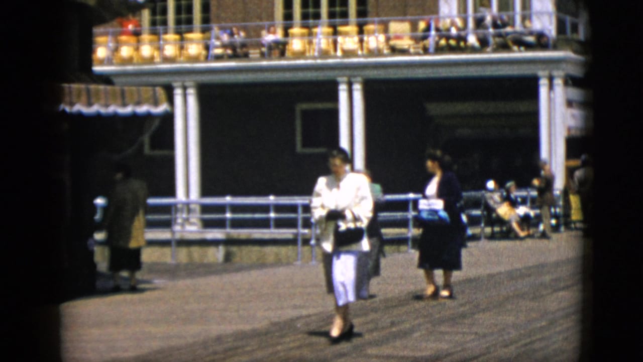 Two women in early-1950s dress walk a sunlit promenade beside a columned building with an upper terrace, attributed to Quebec, Canada, circa 1952.