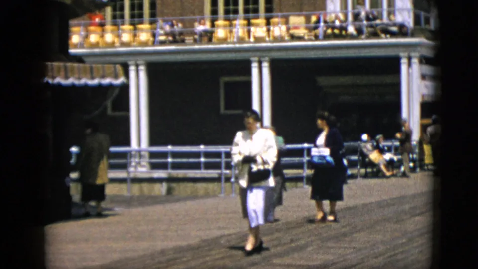 Two women in early-1950s dress walk a sunlit promenade beside a columned building with an upper terrace, attributed to Quebec, Canada, circa 1952.