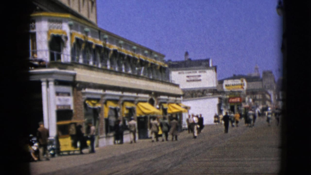 A long commercial building with yellow scalloped awnings and white columns lines a sunlit promenade crowded with mid-century pedestrians; red-and-white signage visible at far right, circa 1952.