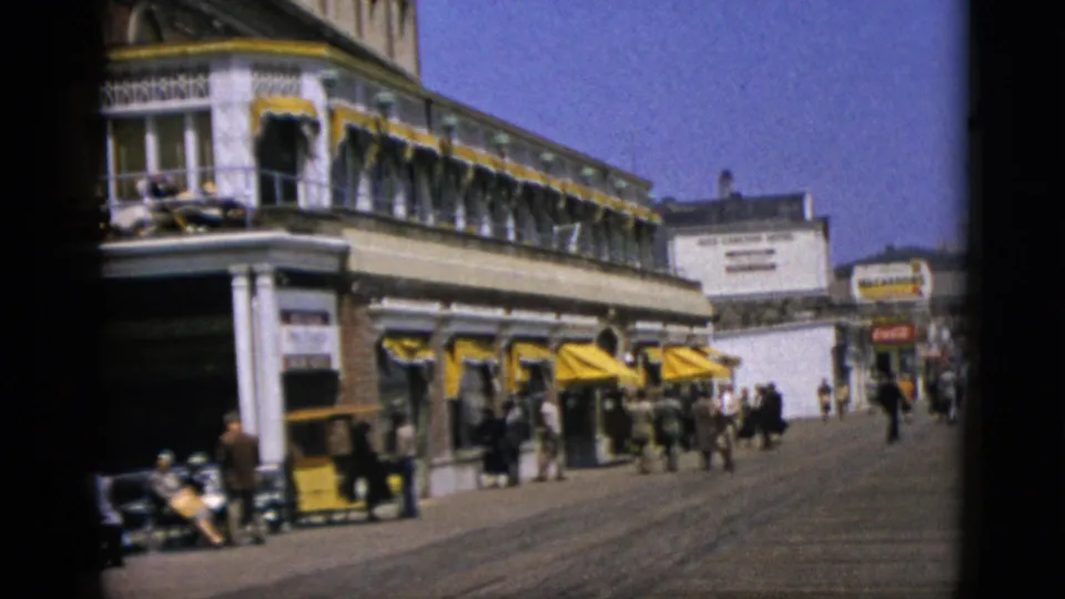 Pedestrians in 1950s dress walk a wide sunny promenade beside a multi-story building with yellow awnings and classical white columns; partial signage legible on facade, circa 1952.