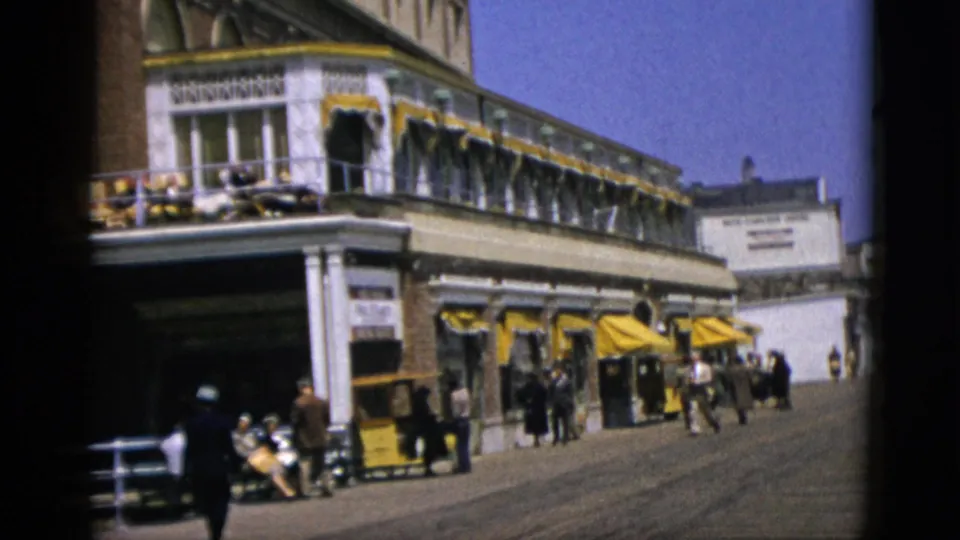 A lone figure walks toward camera on a wide sunny promenade flanked by a long yellow-awninged commercial building; other pedestrians visible beyond, circa 1952.