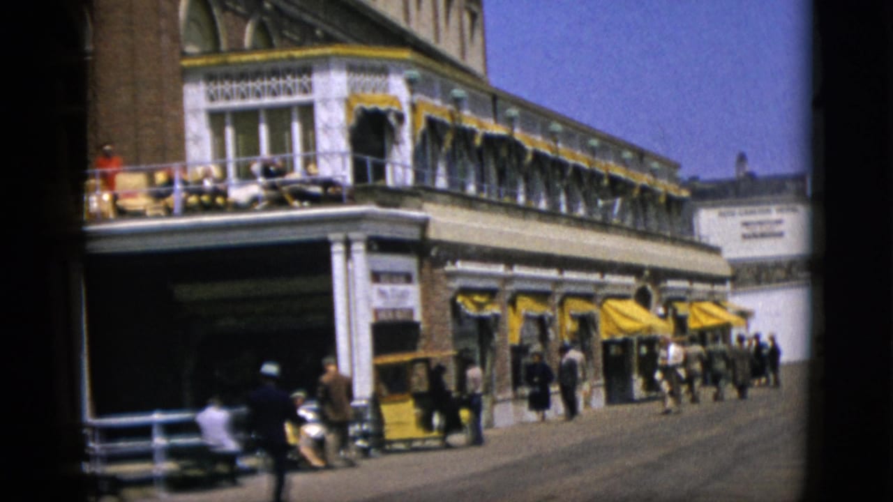 Street-level view of a large red-brick building with yellow awnings and balcony furniture; pedestrians walk on the sidewalk below.