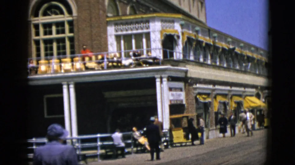 1952 Quebec, Canada: 8mm frame slightly wider; brick building with balcony seating and yellow awnings; a figure in dark clothing in the right foreground.