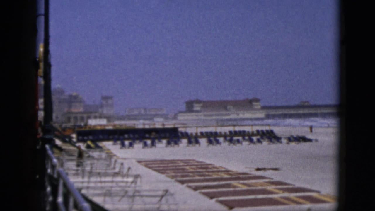 Wide elevated beach view showing rows of rental chairs in mid-ground, rectangular striped platform sections in foreground, and a low-rise beachfront building complex at the waterline.