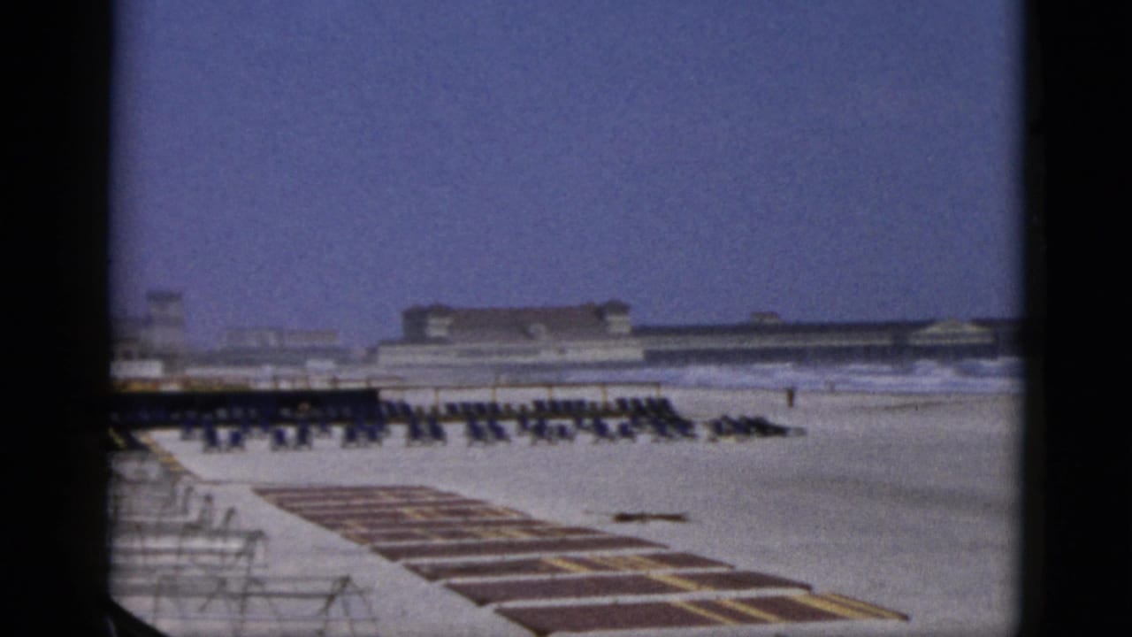 Static wide beach view nearly identical to preceding frame, with rows of chairs in mid-ground, striped platform sections in foreground, and the beachfront building complex at the horizon.