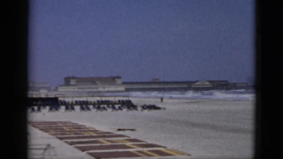 Mid-frame view of a 1950s beach attributed to Quebec, Canada — rows of dark chairs on sand, visible wave action at the waterline, and a beachfront building complex under an open blue-cast sky.