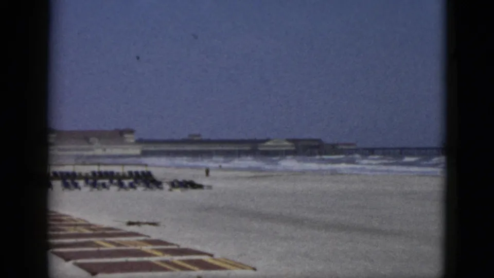 Closing frame of 1950s beach footage attributed to Quebec, Canada, c. 1952 — static wide view of organized chair rows, beachfront facility, and the blue-purple sky cast characteristic of aged 8mm color film.
