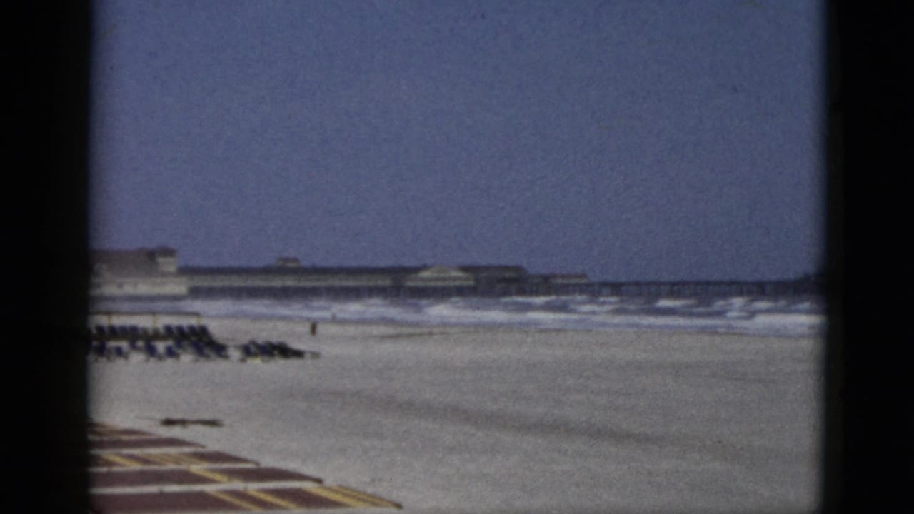 1950s 8mm home-movie frame of a beach attributed to Quebec, Canada: flat sand, gentle surf, striped foreground mat, and a long low beachfront structure at the horizon.