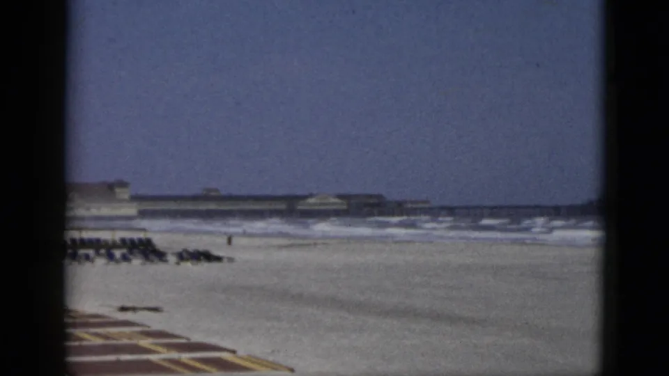 1950s 8mm home-movie frame of a beach attributed to Quebec, Canada: flat sand, gentle surf, striped foreground mat, and a long low beachfront structure at the horizon.