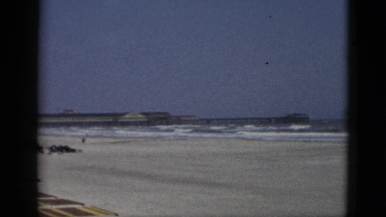 1950s 8mm frame of a beach attributed to Quebec, Canada: wide sandy shore, rolling surf, striped foreground element, and long low structure at the horizon.