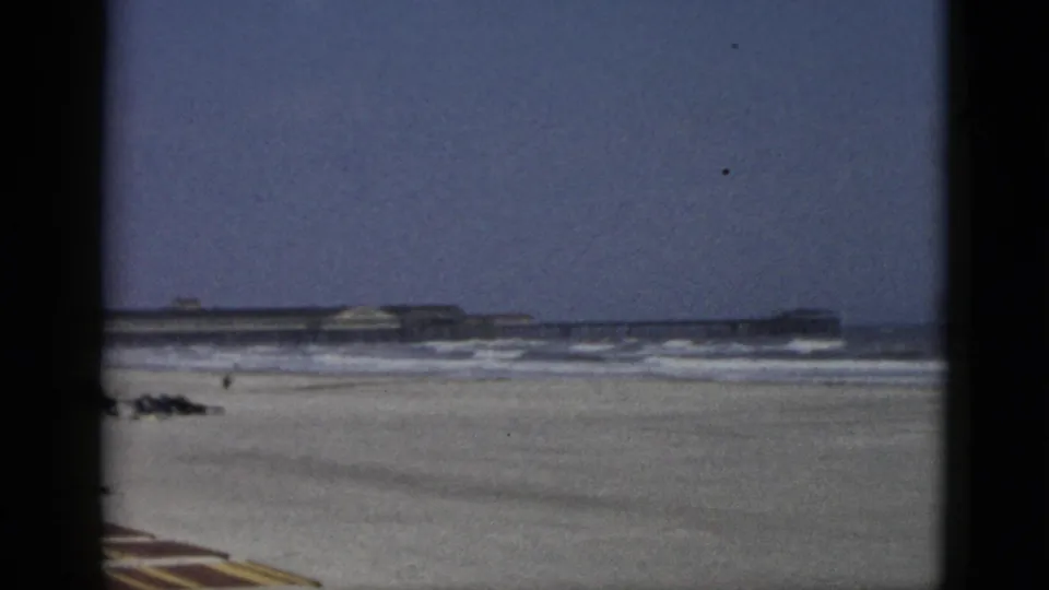 1950s 8mm frame of a beach attributed to Quebec, Canada: wide sandy shore, rolling surf, striped foreground element, and long low structure at the horizon.