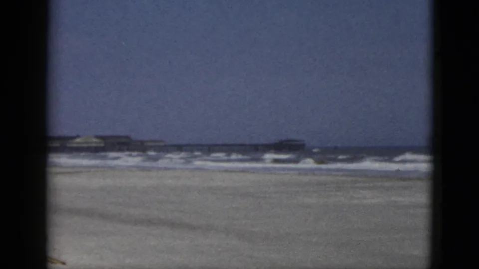 1950s 8mm frame of a Quebec, Canada beach: expanded pale blue sky, surf breaking on sand, and a low beachfront structure on the horizon.
