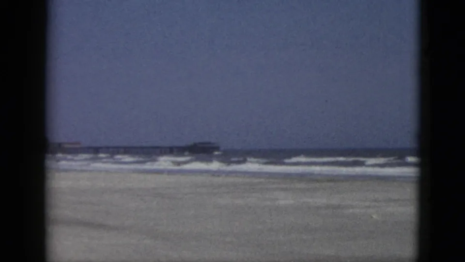 1950s 8mm frame of a Quebec, Canada beach: pale blue sky dominates, with surf, flat sand, and a distant low beachfront structure below.