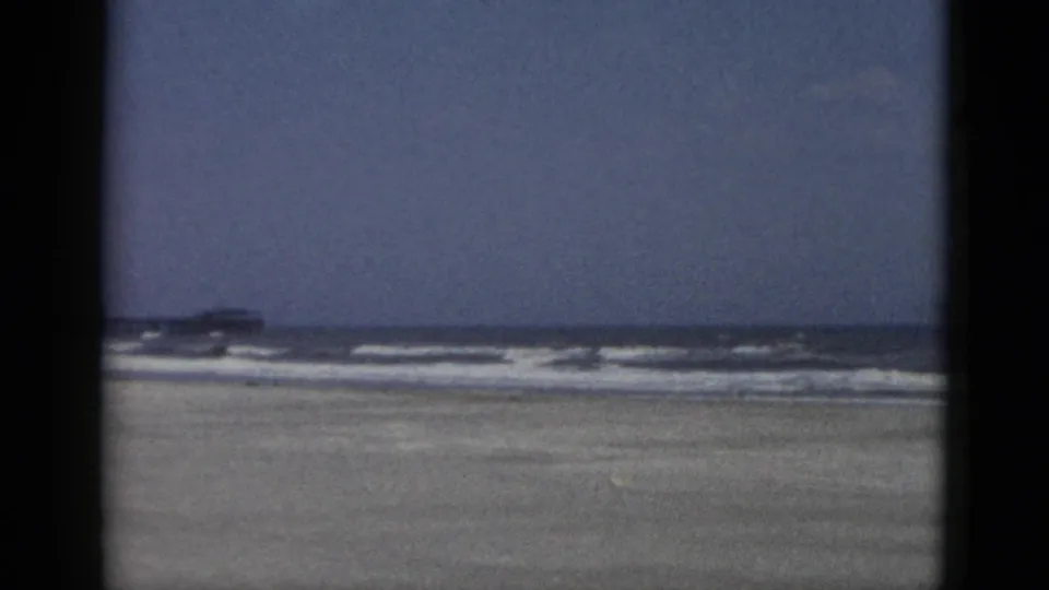 Wide 8mm color view of open sandy beach and surf with a distant pier-like structure at the horizon, Quebec, Canada, circa 1952.