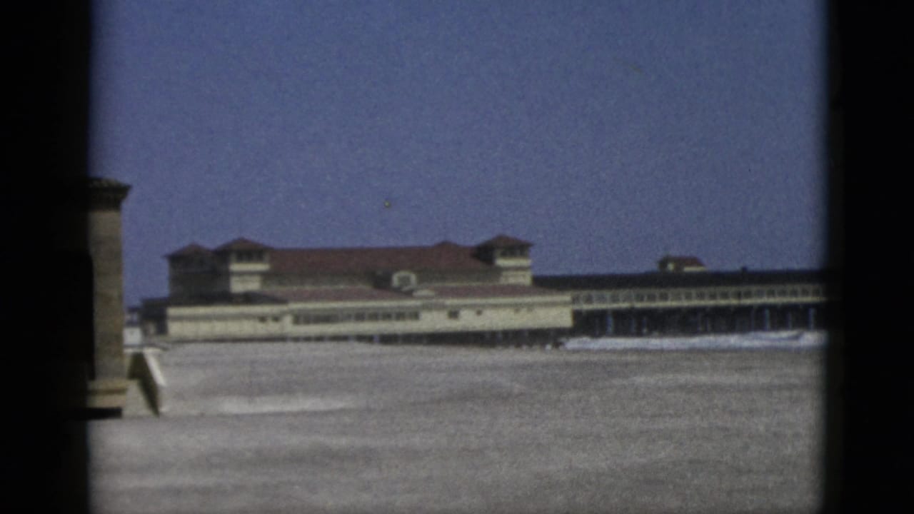 Large seaside building with red tile roof and rooftop towers seen across a sandy beach, framed by dark window bars.