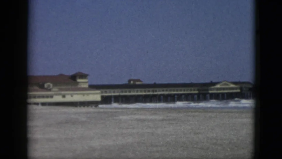 8mm home movie frame showing a large pier-mounted building with visible pilings and sandy beach foreground, likely a Canadian seaside resort circa 1952.