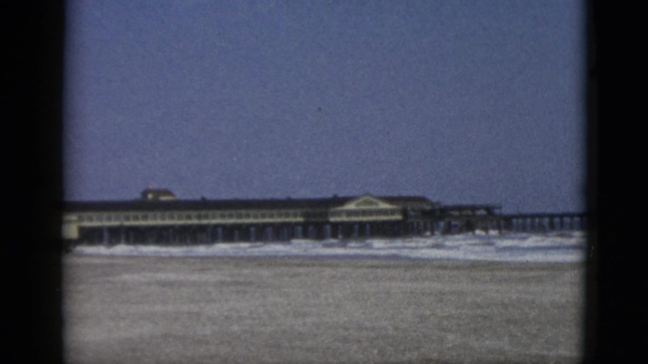 8mm home-movie frame, circa 1952, showing a wide Quebec coastal beach with a wooden pier and mid-century pavilion building in the background.
